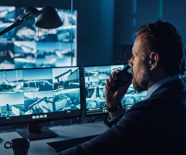 Male Officer Works on a Computer with Surveillance CCTV Video in a Harbour Monitoring Center with Multiple Cameras on a Big Digital Screen. Employee Uses Radio to Give an Order or Report.